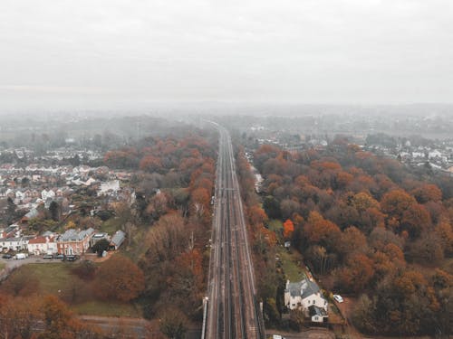 Breathtaking aerial view of railway road going through lush autumn trees near city residential district with various cottages against gloomy sky