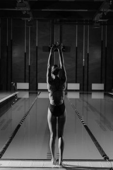 A woman in a swimsuit stretches at the poolside in a monochrome indoor setting.