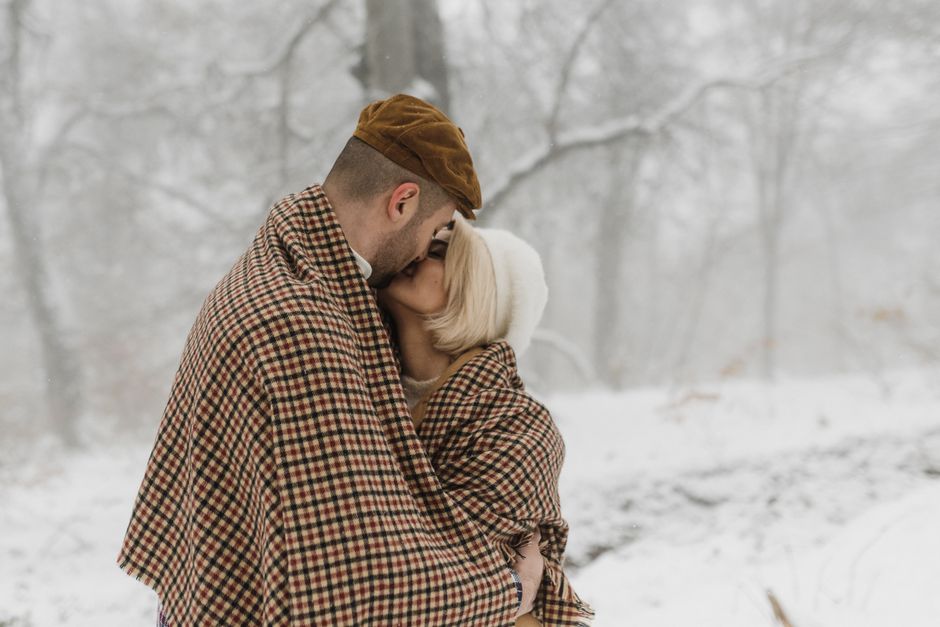 A loving couple wrapped in a blanket, sharing a kiss in a snowy winter forest.