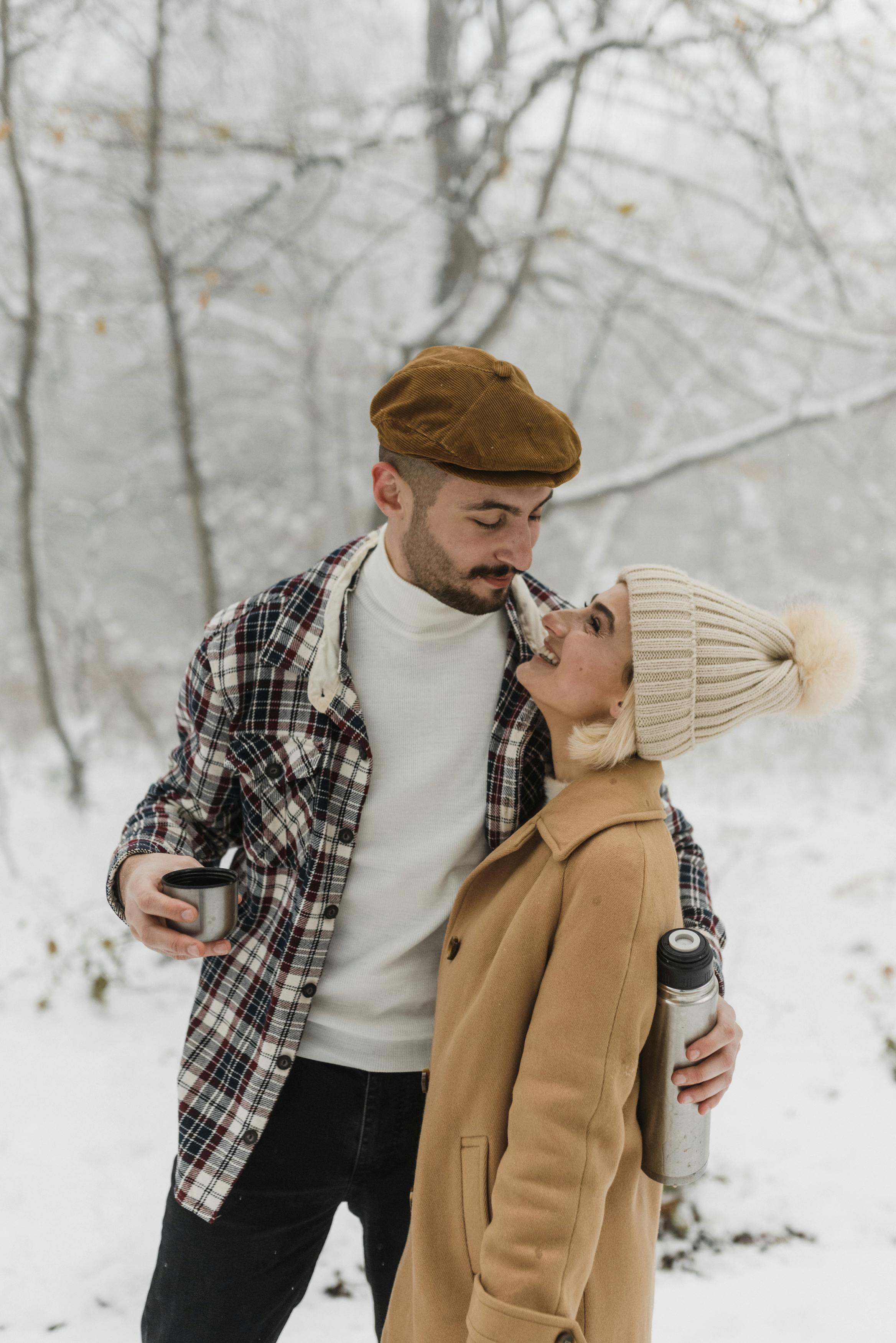 Happy couple embracing in snowy forest, sharing a warm beverage during a cozy winter day.