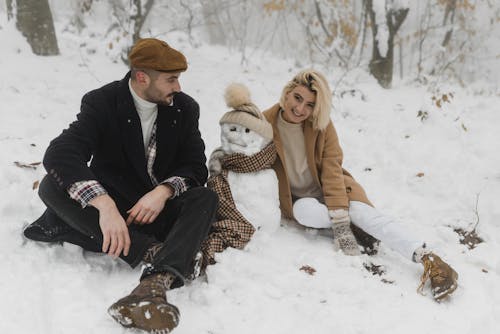 Couple Sitting on Snow Covered Ground Near the Snowman