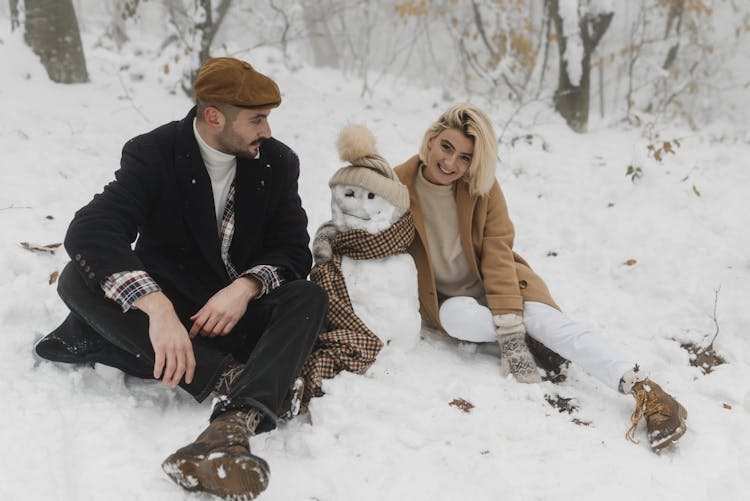 Couple Sitting On Snow Covered Ground Near The Snowman