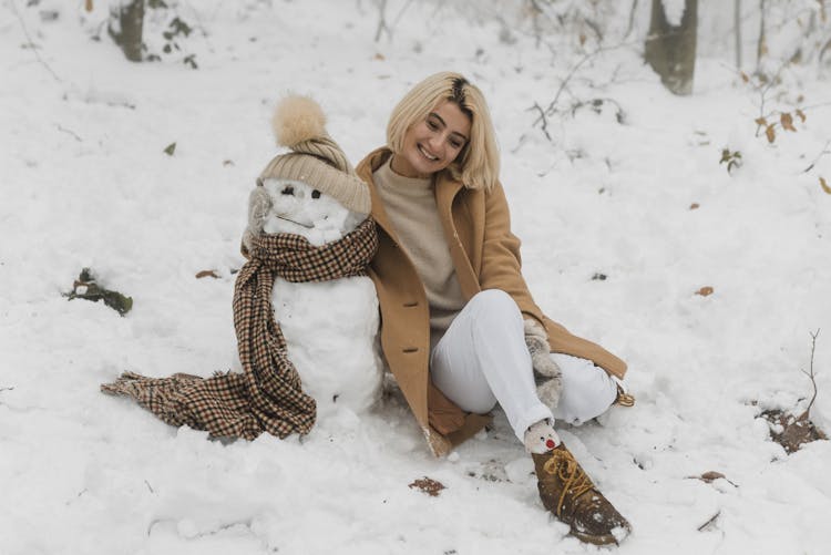 Happy Woman Sitting On Snow Covered Beside The Snowman