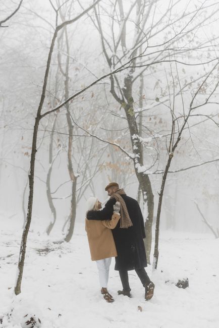 A couple walks through a serene snowy forest, embracing cold winter weather in warm clothes.