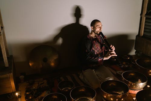 A Man Playing Flute while Sitting on the Ground Surrounded by Singing Bowls