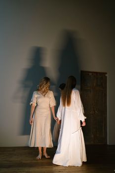 Three women in white dresses holding hands, casting long shadows on a wall.