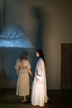 Two women standing in a dimly lit room, casting shadows on the wall, creating an artistic silhouette.