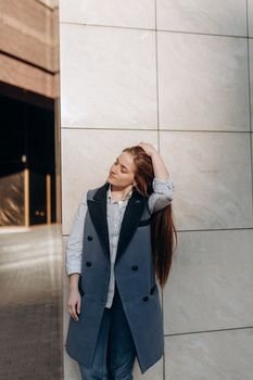 Young thoughtful female with eyes closed in stylish outfit pondering near wall with tile on street of city