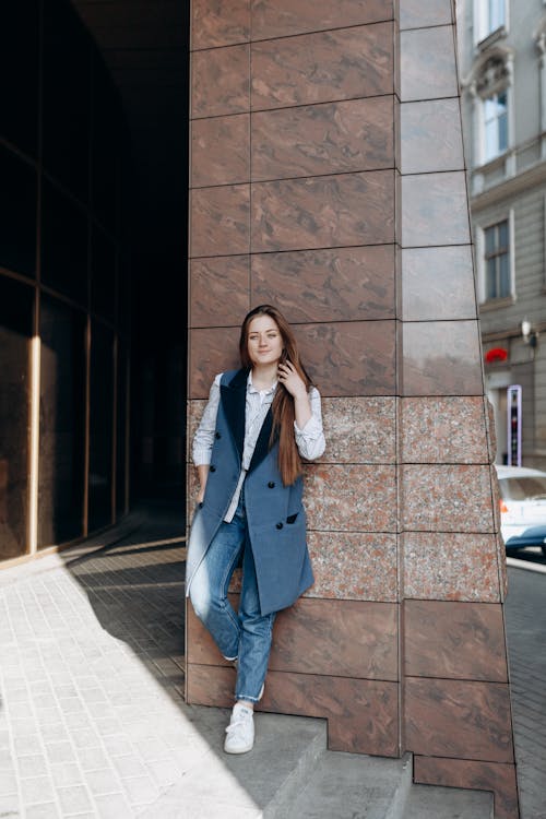 Full body cheerful female smiling and looking at camera while leaning on wall of contemporary building