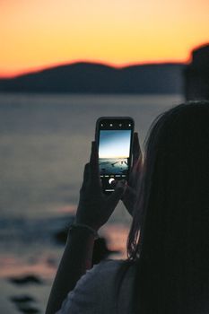 A woman photographs a stunning sunset over the sea using a smartphone, highlighting technology in scenic views.
