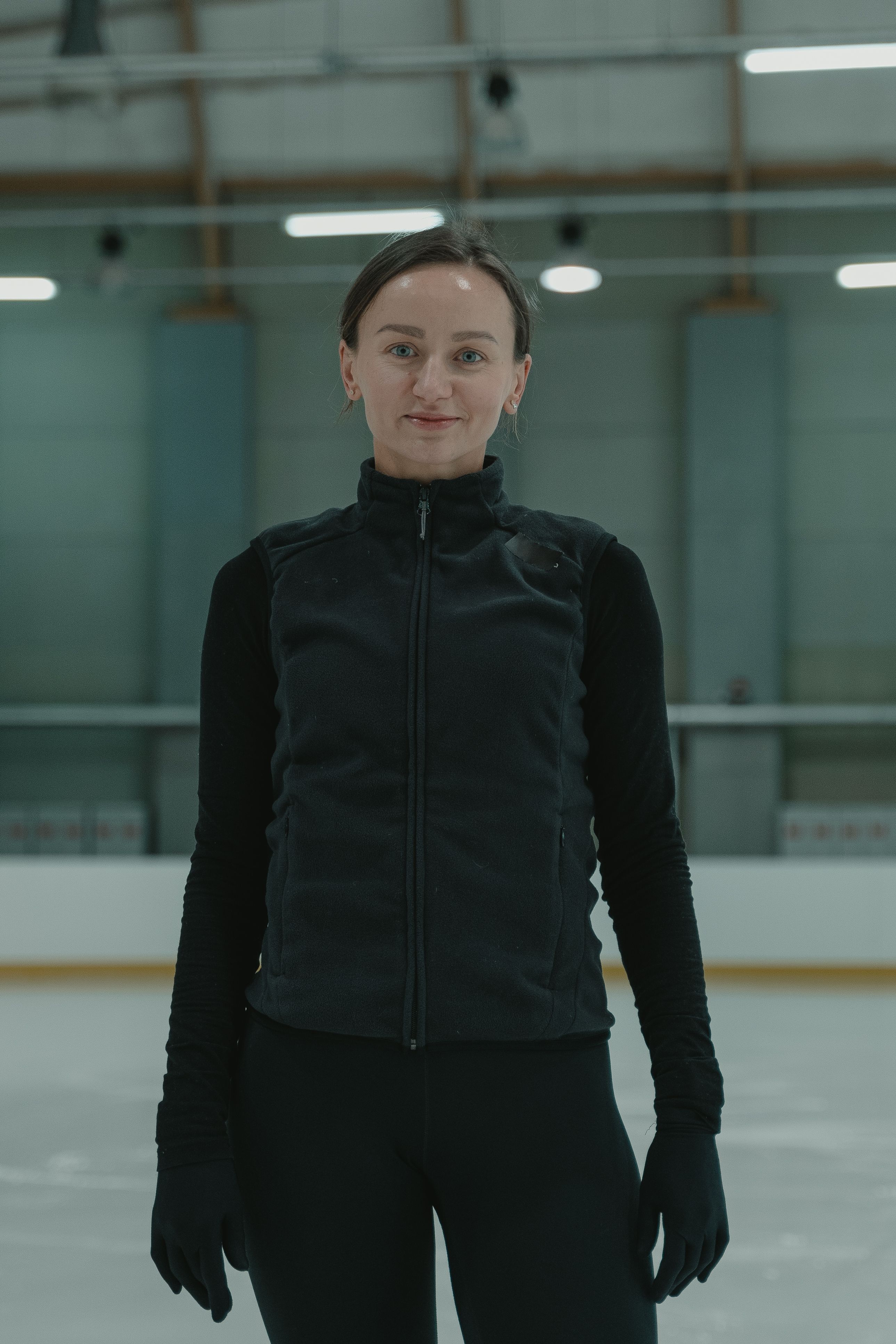 Woman wearing a black jacket poses confidently on an indoor ice rink, ready for skating.