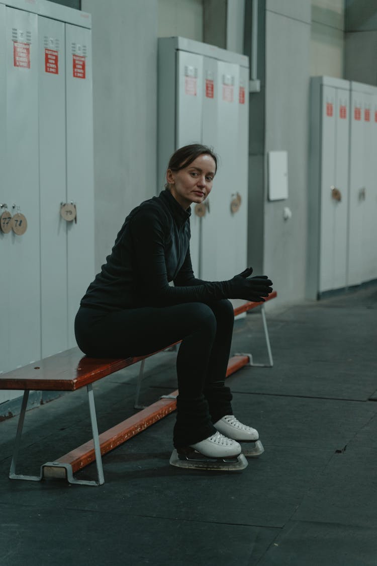 Woman In Black Clothes Wearing Ice Skating Shoes Sitting On Bench 