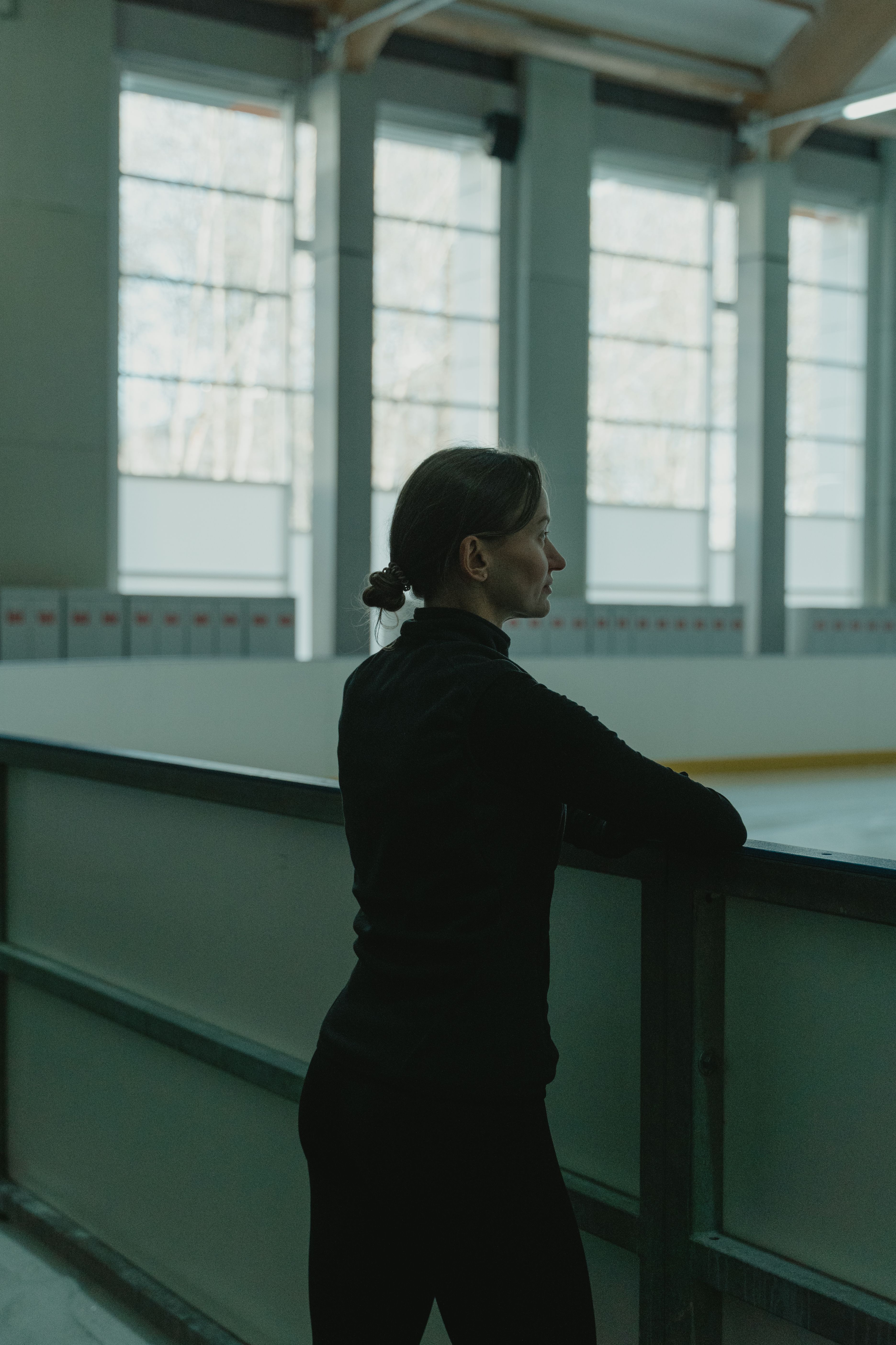 A woman in black clothing stands thoughtfully by an ice rink indoors.