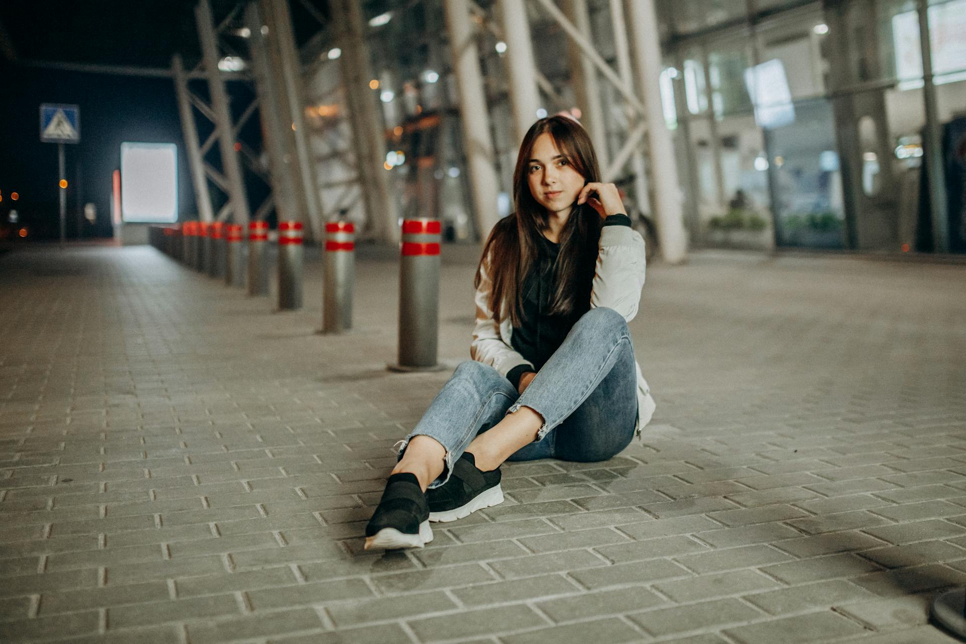 Full body young female in denim resting on concrete tile near fence posts on blurred background of modern building