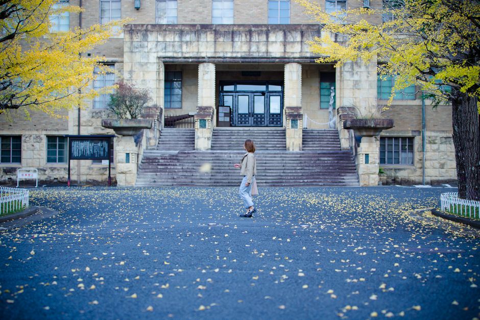 女性が秋の木々と落ちた黄色い葉に囲まれた歴史的な建物の前を散策しています。