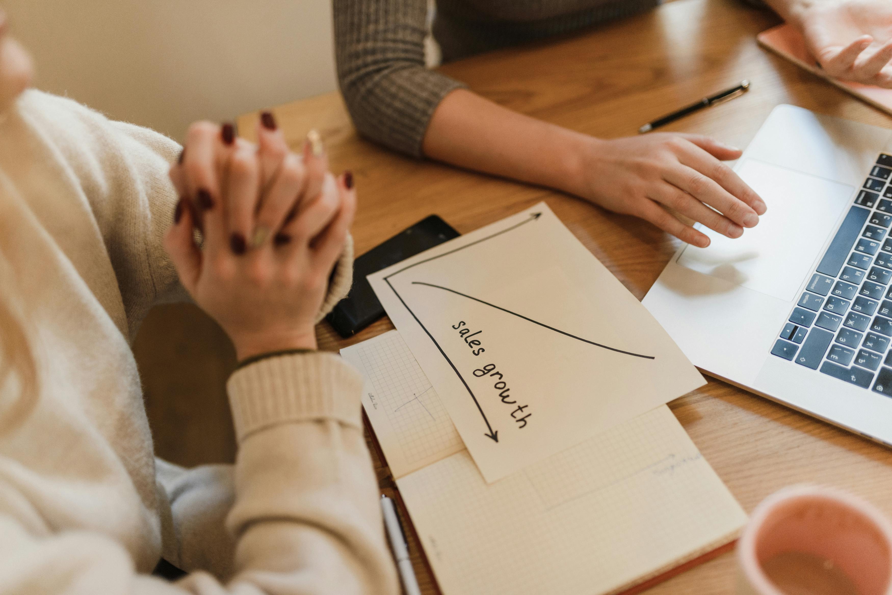 Two women collaborate at a desk discussing sales growth with a laptop and notes.