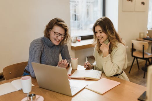 Two women laughing and working together in a cozy office setting with laptops and notebooks.