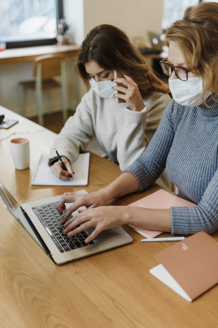 Women In Face Masks Sitting At Desk Working On Laptop