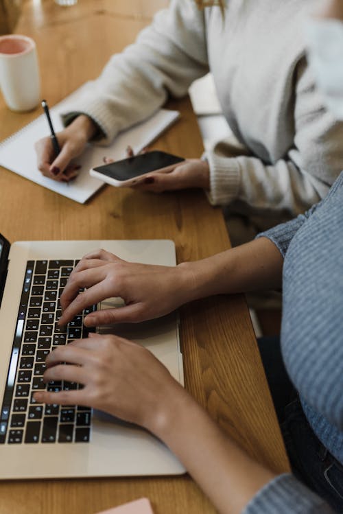 Person in Gray Sweater Typing on a Laptop Beside a Person Writing on Notepad while Holding a Cellphone