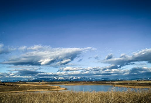 Tranquil view of a vast landscape with a pond and distant mountains under a cloud-filled blue sky.