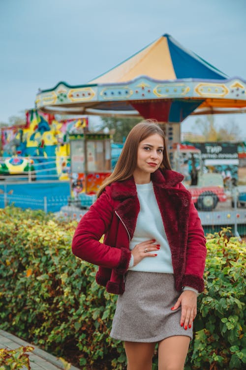 Attractive brown haired female in casual clothes standing against carousel and holding hand on waist while looking at camera