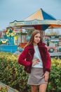 Attractive brown haired female in casual clothes standing against carousel and holding hand on waist while looking at camera