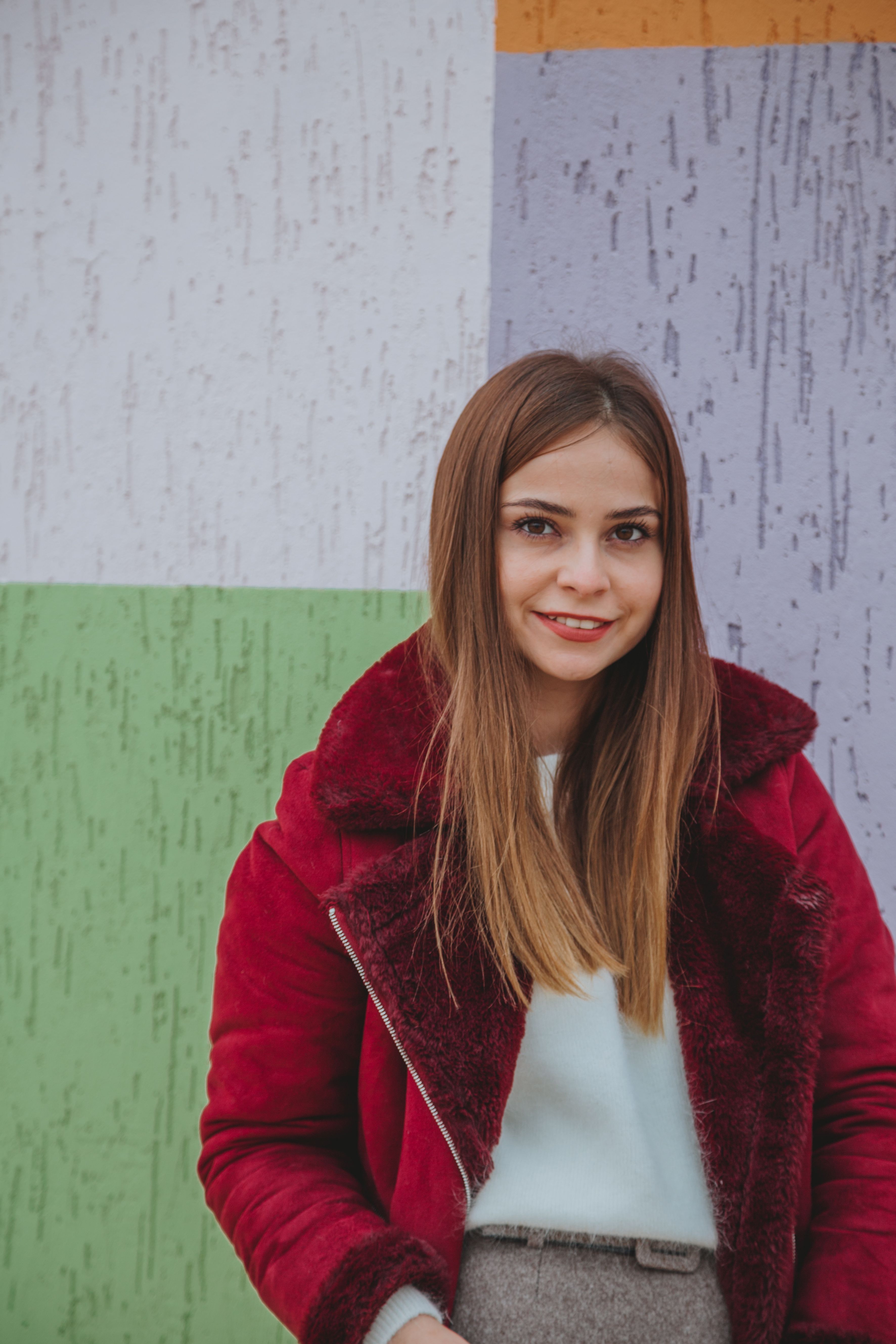 Young pretty female in trendy jacket standing near concrete multicolored building and looking at camera
