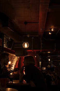 Side view of couple chatting while resting in dark building of old pub with bright lamps
