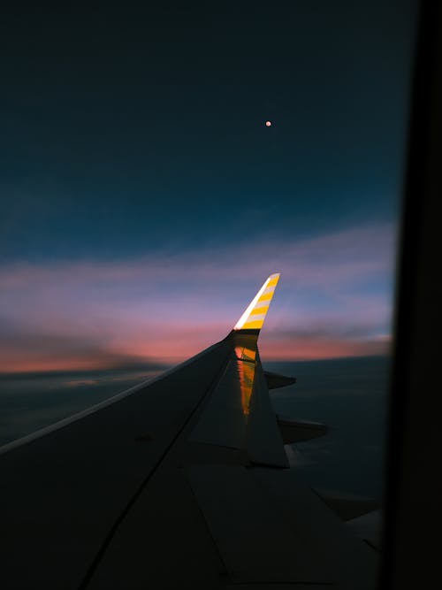 Through window of wing of modern aircraft flying over fluffy clouds in dark colorful sunset sky