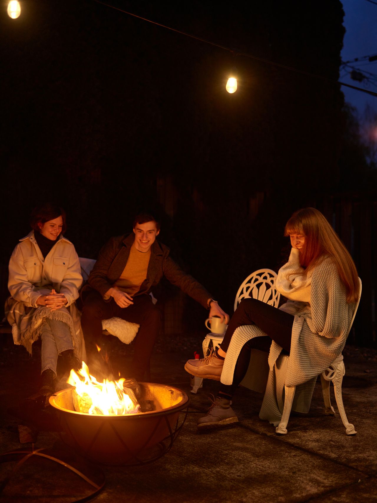 Friends wrapped in warm blankets and knits sitting around a glowing fire bowl on a small patio at night, with a mug waiting on a side stool