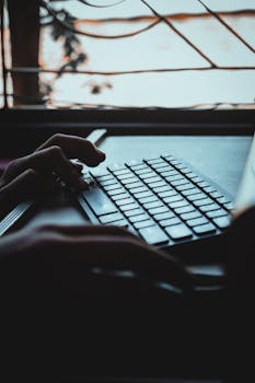 Close-up of hands typing on a laptop by a window with a nature view