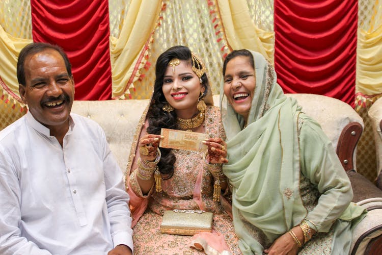 A Woman In Traditional Wedding Dress Holding Paper Money While Sitting In Between Her Parents