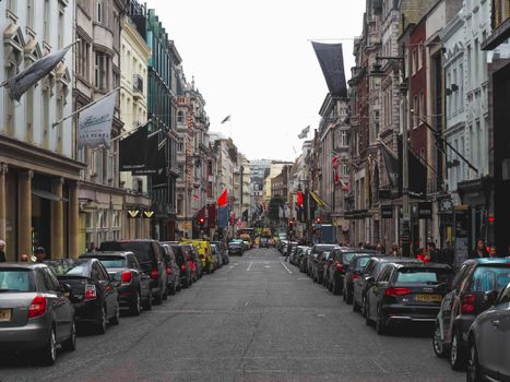 A busy street lined with parked cars and bustling shops in central London, UK.
