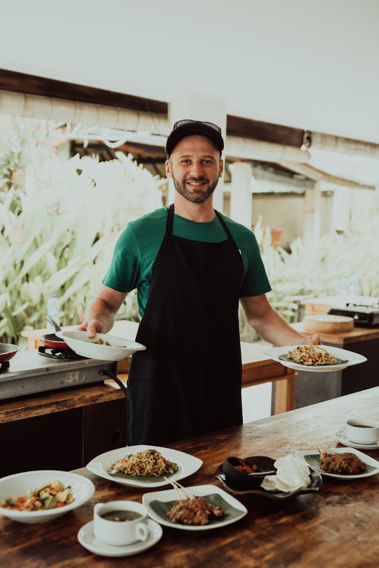 A Man In Green Shirt Wearing Apron Holding Plates With Food