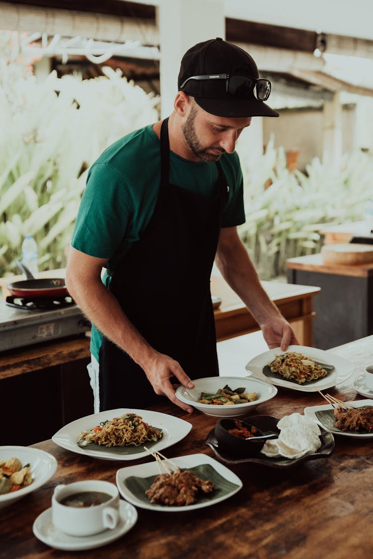 A Man In Green Shirt Wearing Apron Holding Plates With Food
