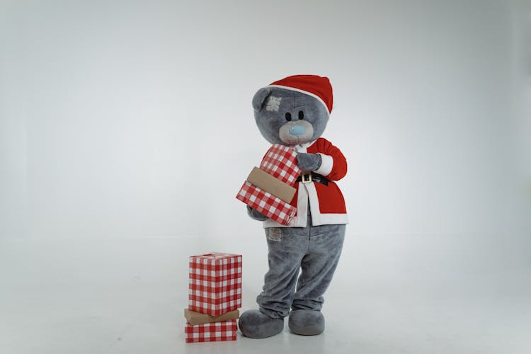 A Teddy Bear Mascot In Santa Claus Costume Holding Christmas Presents