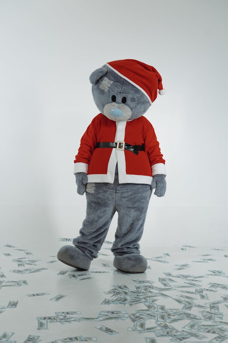 A Teddy Bear Mascot In Santa Claus Costume Looking At The Cash Money On The Floor