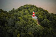 Aerial view of red and white lighthouse located on hilltop among high green trees and near calm ocean in daytime