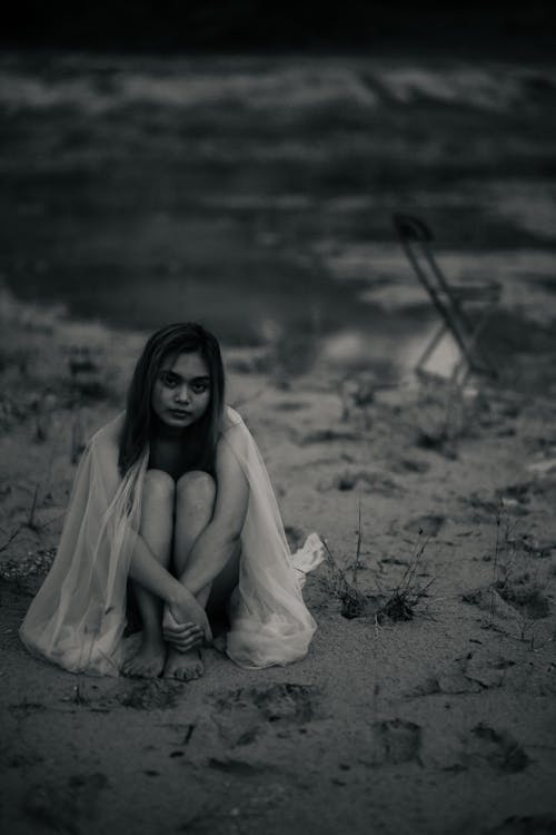 Black and white of young barefoot ethnic female in transparent fabric sitting on coast against pond and looking at camera