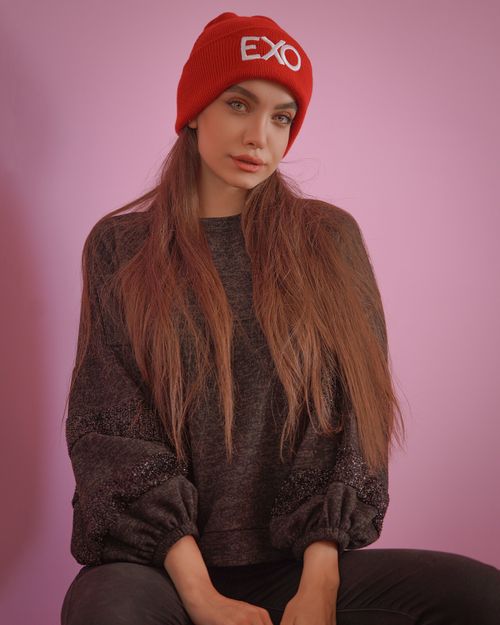 Free Portrait of a woman in a red EXO beanie, long hair, and sweater against a pink backdrop. Stock Photo