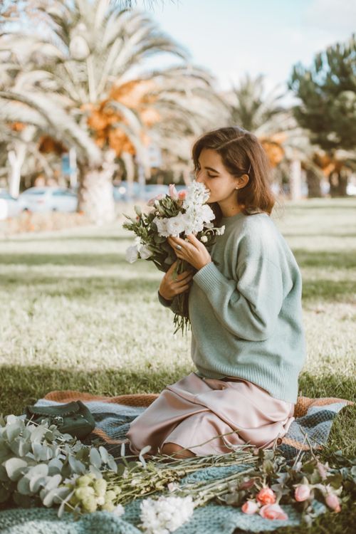Free Young woman sits on a picnic blanket, savoring a fresh bouquet of flowers in a sunny park. Stock Photo