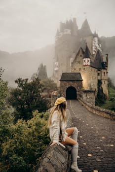 A woman sits on a bridge leading to the fog-covered Eltz Castle in Germany.