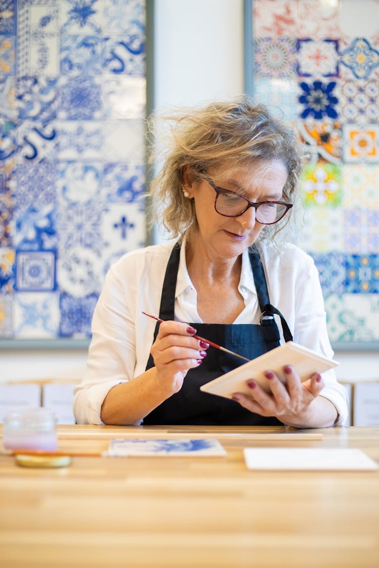 A Woman Wearing Eyeglasses Painting A Ceramic Tile