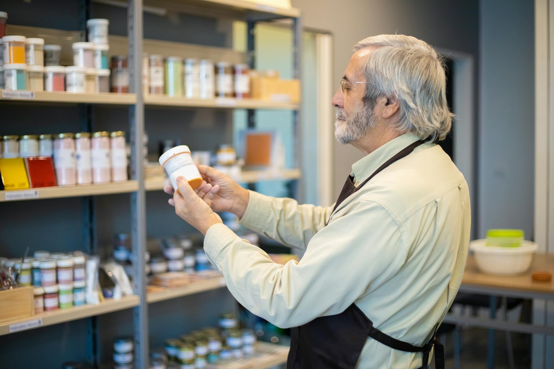 Senior man in apron organizing paint jars on a store shelf indoors.