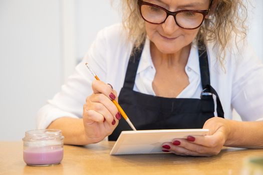 Close-up of a senior woman painting a ceramic tile, focusing on creativity and detail.