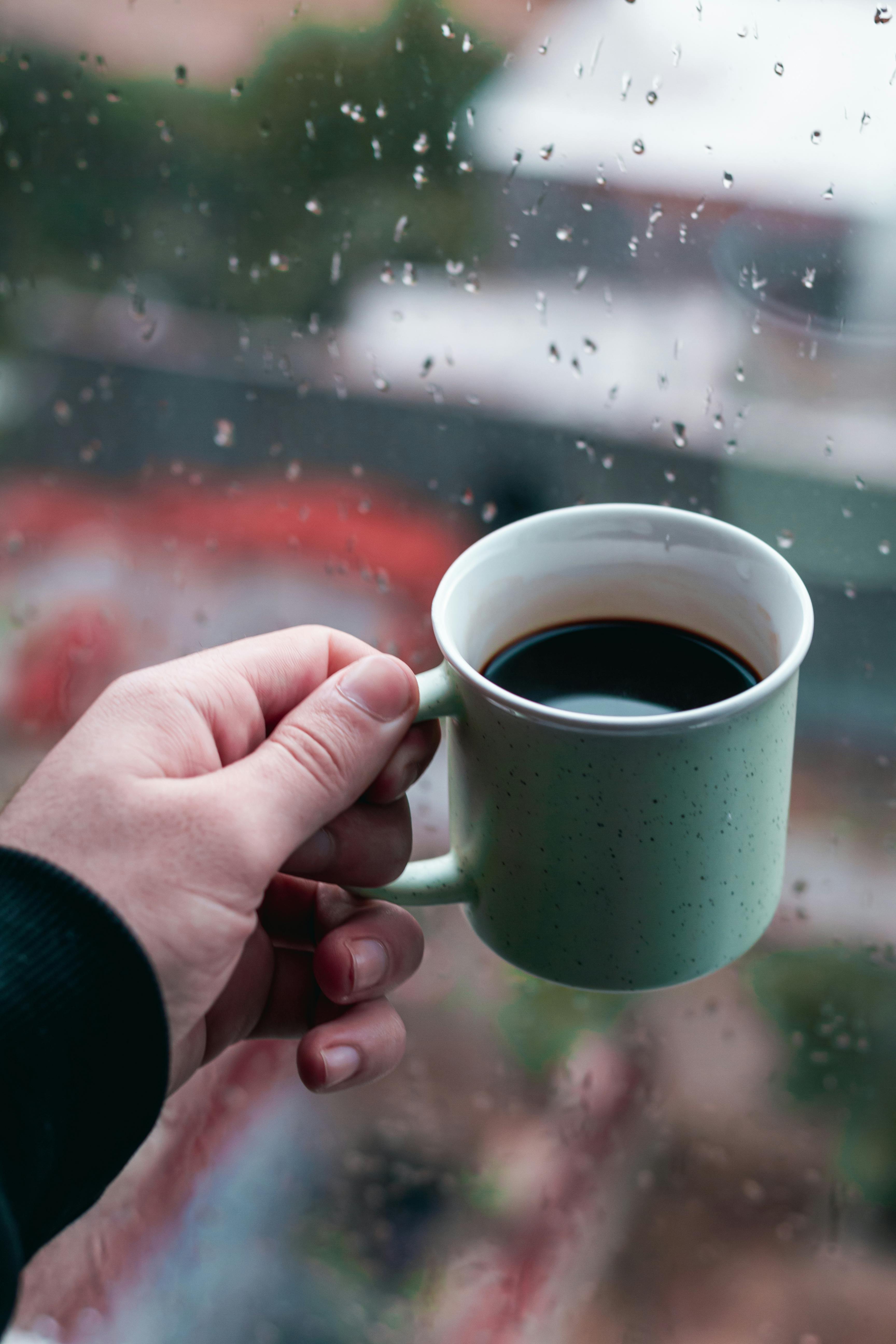 Free A person holds a cup of black coffee indoors, gazing out a rain-specked window. Stock Photo