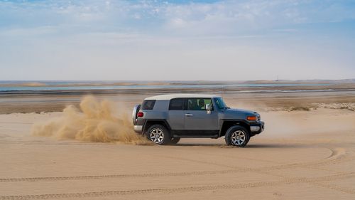 Free Side view of anonymous traveler driving contemporary off road car drifting in desert near sea under cloudy blue sky Stock Photo
