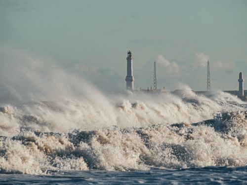 Free Powerful ocean waves crash near a lighthouse under a clear sky, showcasing nature's force. Stock Photo