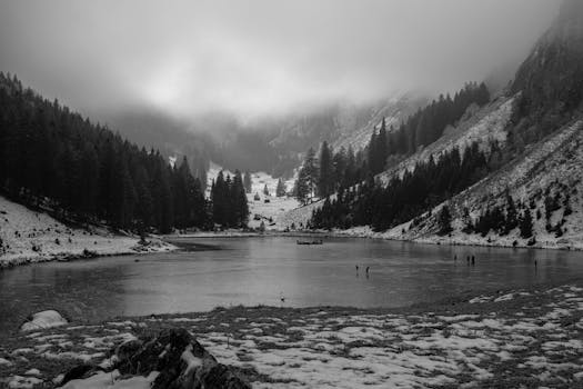 A tranquil winter scene with a misty lake surrounded by snowy mountains and trees.