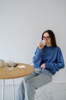 Young female in casual wear with cup of hot drink sitting in armchair and looking away in building